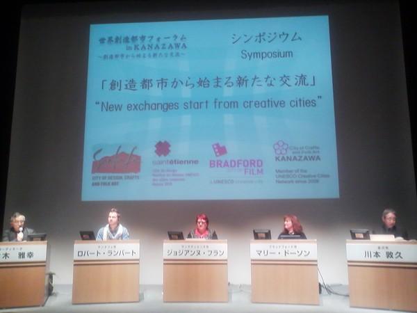 プロジェクトスクリーンの前の用意された席に座る外国人男女3名とスーツを着た男性2名の写真