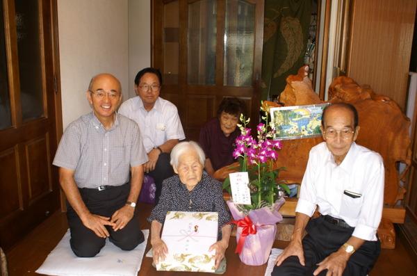 送られた花の横に座り、記念品を持つ大前 すまゑさんと、ご家族、関係者と市長の写真