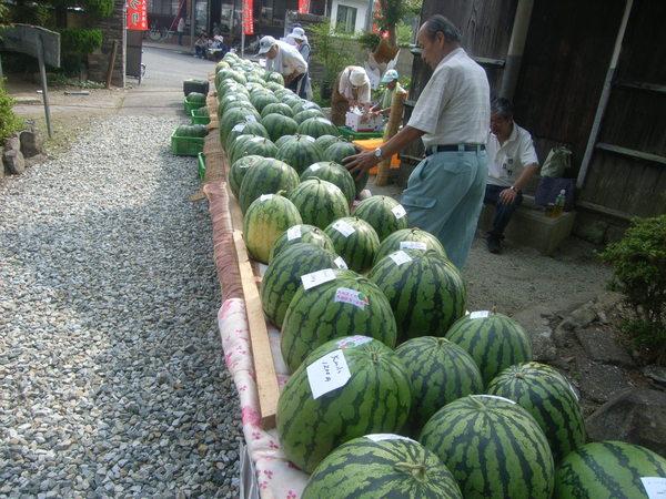 男性が、台の上に並べられた沢山のスイカの中の一つを実際に手で触って見ており、奥に写っている男性もスイカの品定めをしている様子の写真