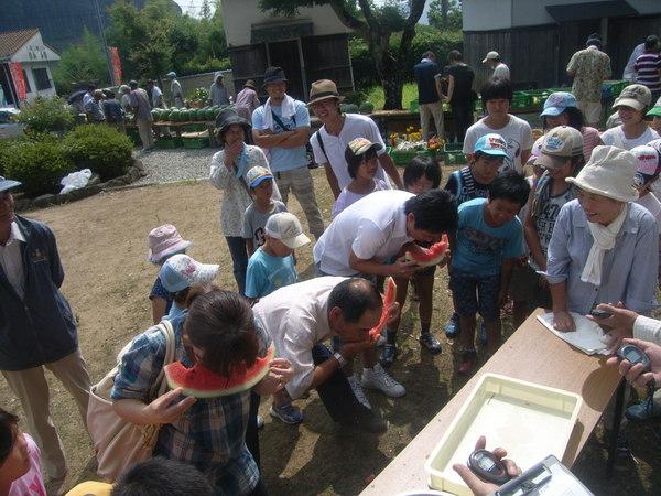 スイカの早食い競争で男性2名と女性1名が競って食べているのを、周りの人達が覗き込んでみている様子の写真