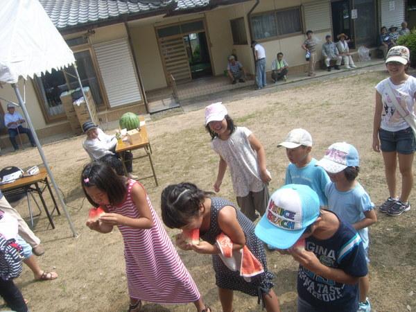 女の子3人がスイカの早食い競争でスイカにかぶりついており、後の子ども達も笑顔で見ている様子の写真