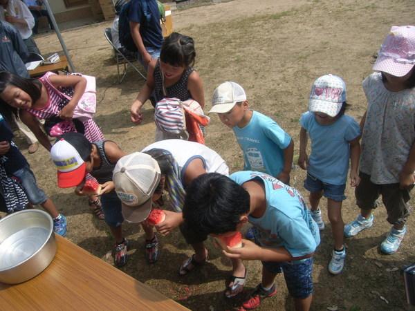 男の子3人がスイカの早食い競争に参加しスイカにかぶりついており、周りの子どもたちが誰が早いか覗き込んでいる様子の写真