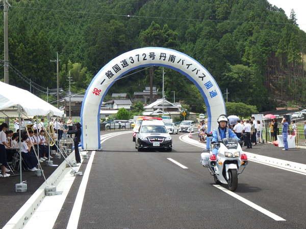 丹南バイパスを白バイ隊員を先頭にパトカーや乗用車が走っているのを沿道の人々が見ている写真