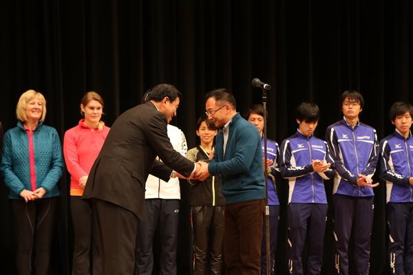 舞台で主催者の男性と男性選手が両手で握手しているのをジャージ姿の選手らが見て拍手している写真