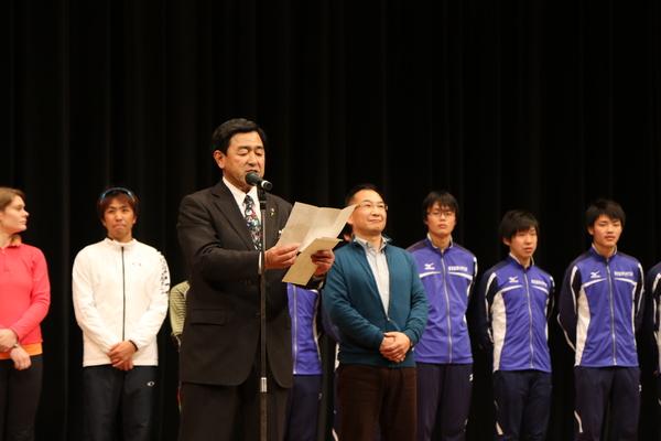 舞台で挨拶をしている男性の後ろでジャージ姿で並んで聞いている選手らの写真