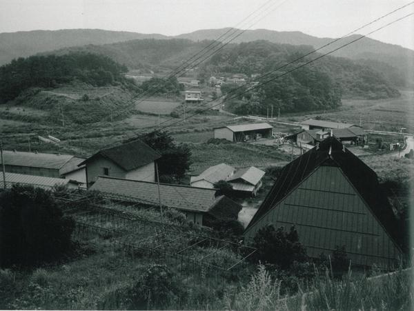 奥には山が連なっており、人影のない村で田んぼと住宅や倉庫が写っている白黒の写真