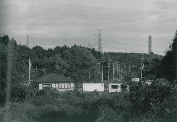 山の向こうに送電鉄塔がいくつか立っており、中央に住宅と倉庫が並び、手前には草が生い茂っている様子の白黒の写真