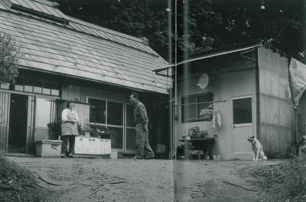 住宅の庭先を歩いている佐藤 義明さんとにエプロンを付けたひろこさんが写っており、倉庫の前に一匹の首輪を付けた犬お座りをしている白黒の写真