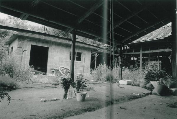 納屋の屋根の下にペットボトルに菊の花束が生けられており、建物の周りには雑草が生い茂って雑然とした風景の白黒の写真