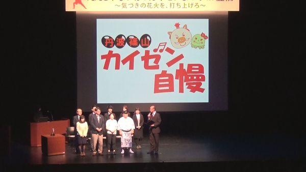 会場のスクリーンに「丹波篠山市 カイゼン自慢」とのど自慢風に文字が書かれており、舞台の上ではマイクを持っている男性が浴衣を着た男性やスーツを着た男女、黄色いカーデガンを着た女性などに、話をしている様子の写真