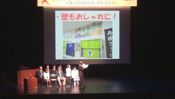 会場のスクリーンに「壁もおしゃれ」と書かれ、窓口を工夫している様子の写真が映し出されており、映像を見ながら、担当者が説明をしている様子の写真