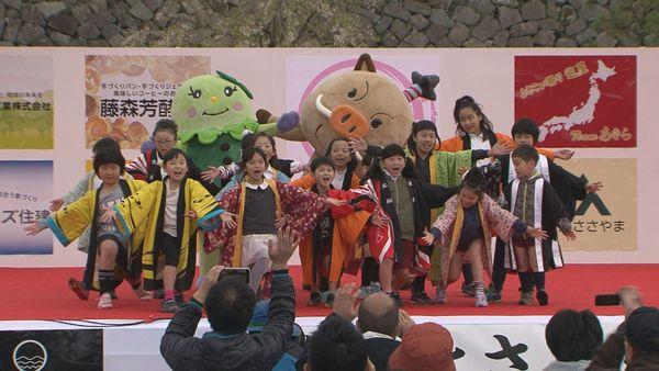 舞台の上で着ぐるみを来た人2人と法被を着た子供たちが両手を広げポーズをとっている写真