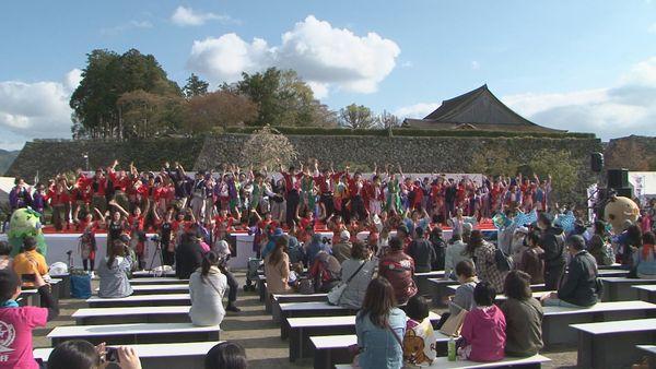 篠山遺跡で、舞台の上と下に、法被を着たたくさんの人たちが両手を挙げているのを、座ってみている見物人の写真