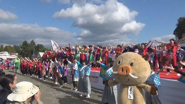 舞台の上と下で色々な衣装を着た人たちと、まるいのが両手を挙げてポーズをとっている写真