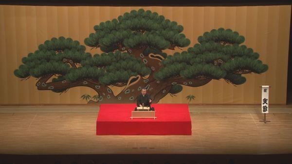舞台の後ろに飾られている絵には大きな盆栽が描かれ、そこで落語を話されている写真