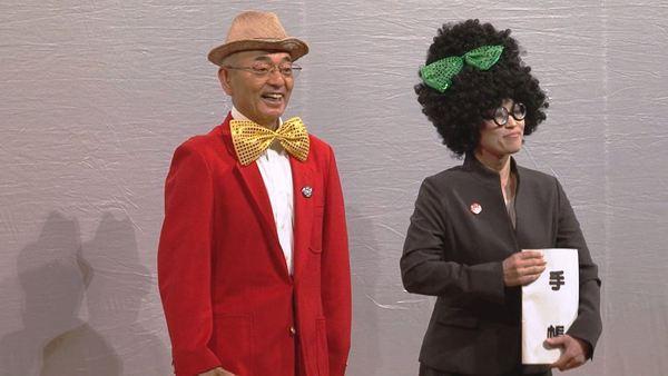 帽子を被り蝶ネクタイに赤いブレザー姿の市長が手帳を持ったパーマヘアーにリボンを付けたスーツ姿の秘書とコントしている写真