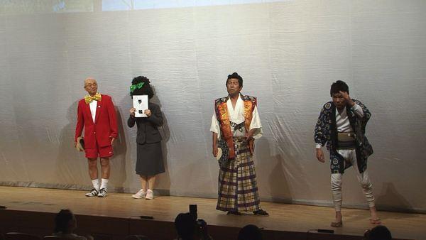 帽子を片手に真っ赤なブレザーと半ズボン姿の市長、パーマヘアーにリボンを付けたスーツ姿の女性が手帳で顔を覆っており、扇子を持った侍とパッチにはんてんを羽織った侍が舞台に立っている写真