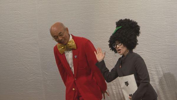 手帳を持ったパーマヘアーにリボンを付けたスーツ姿の秘書が市長のいる右側へ手を上げ、蝶ネクタイに赤いブレザー姿の市長が話をしている写真