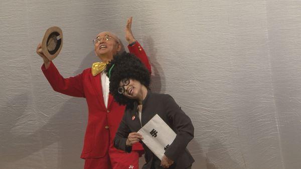 帽子を右手に持った蝶ネクタイに赤いブレザー姿の市長が手を上げて、手帳を持ったパーマヘアーにスーツ姿の秘書が右へ体を傾けている写真