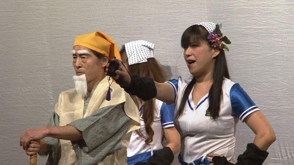 黄門さまの横でチアガール風の衣装に日本タオルを載せた女性が印籠を差し出している写真