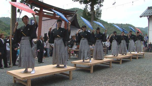 紋付羽織袴を着た男女が舞台の上で踊り傘をを手に持ちポーズしている写真