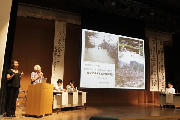 大芋地区活性化委員会代表の江坂 道雄さんが「大芋では水害と土砂災害」の映像を見ながら講演している写真