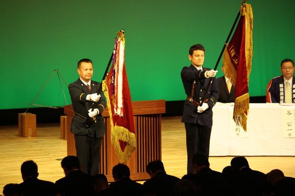 壇上で制服姿の男性2名が客席側に向けて旗を持って立っている写真