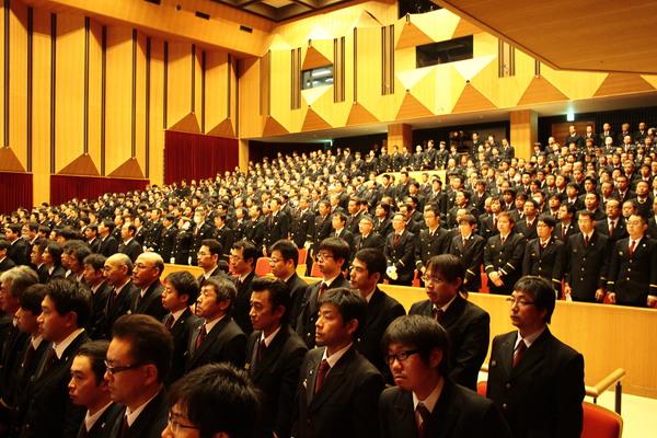 出初式にてスーツ姿や制服姿の男性らが客席に立っている写真