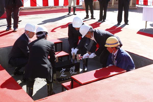 ヘルメットをかぶりスーツを着た男性4名が「定礎」と書かれている礎石を据えるために、紅白のリボンで巻かれた木槌を持って、礎石の四隅を叩いている様子の写真