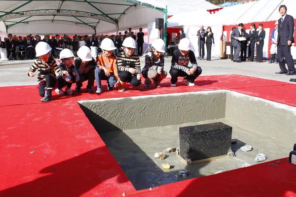 ヘルメットをかぶった男子と女子の児童7名が、四角に囲まれてた赤い絨毯の上にかがんでおり、その前には礎石が据えられ、礎石の周りには子ども達の夢が書かれてた石が置かれている様子の写真