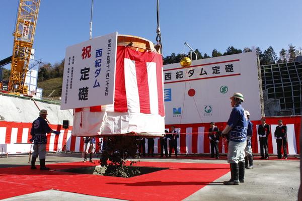 祝 世紀ダム 定礎と書かれた看板の後ろに大きな紅白の幕に覆われた樽のようなものがあり、四角い枠の中に、樽のようなものからセメントが流し込まれている様子の写真