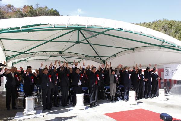 テントの下に胸に胸章を付けた出席者が大勢集まっており、椅子から立ち上がって両手を上げて万歳三唱をしている様子の写真