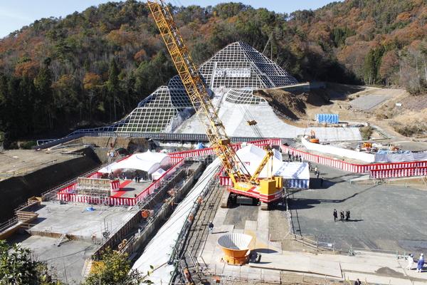 紅白の横断幕が張り巡らされセレモニーが行われている西紀ダムの建設現場全体の様子を山の上の方からうつしている様子の写真