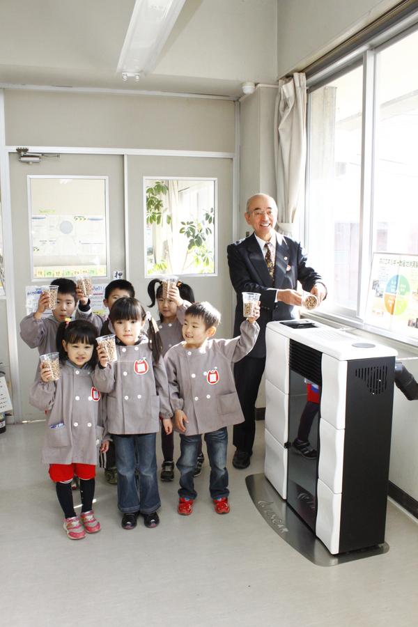 園児6名がプラスチックの透明コップに木質ペレットを入れて持っており、市長が木質ペレットをペレットストーブの燃料入れに入れようとしている様子の写真