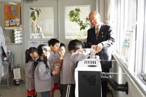 幼児達が設置されているストーブに木質ペレットを入れるのを順番待ちしており、一人がコップに入った木質を入れており、市長が見守りながら手を添えている様子の写真