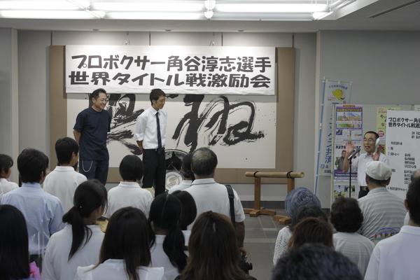 沢山の激励会参加者が見守る中、角谷 淳志選手と男性が、マイクの前に立つ男性を見ている写真