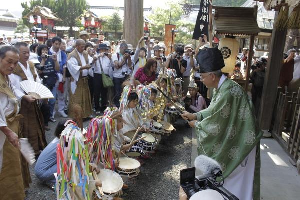 紙飾りをして締太鼓を叩いている子供たちの頭上に神主がお祓いを行っている写真