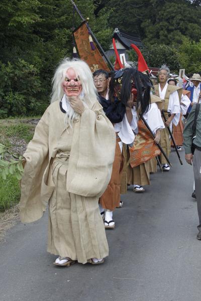 白髪で強面のお面を被った男性を先頭に着物姿の男性たちが旗や獅子舞の頭を手にして1列になって練り歩いている写真