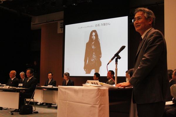 スクリーンにはシンガーソングライター白石 乃梨さんが映っていて、男性が立ってマイクで話をしている写真