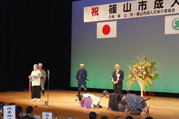 舞台の上で水戸黄門の格好をした男性が立っていて、その横で侍姿の男性達が膝まづいて頭を下げている写真