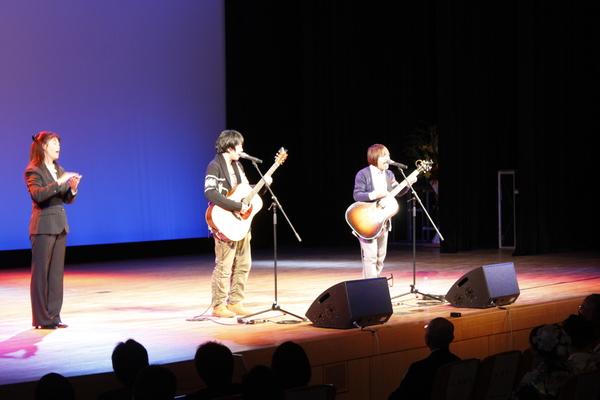 舞台の上でギターを持った男性2人が歌を歌って、その横で手話をしている女性が立っている写真