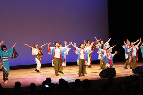 舞台の上で法被姿の人達が10名ほど立っていて、両手を大きく広げて踊っている写真