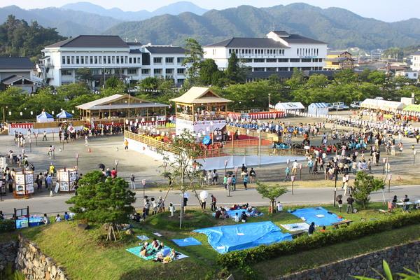 祭り会場の周りに集まる人々や、シートをひいて座っている人々の写真