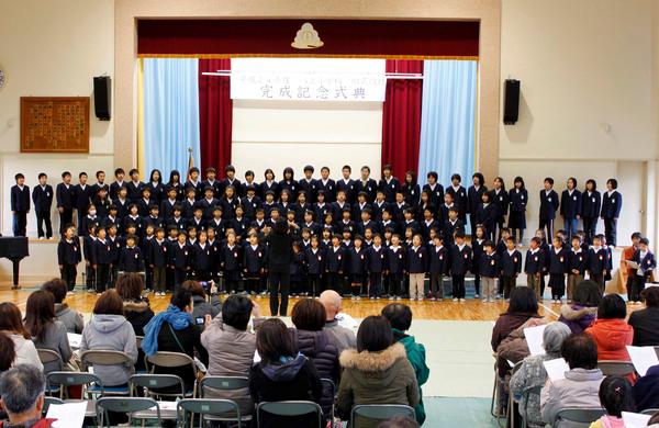 八上小学校の耐震改修完成記念式典にて体育館のステージで生徒らが指揮に合わせて合唱しているのを観客が見ている写真