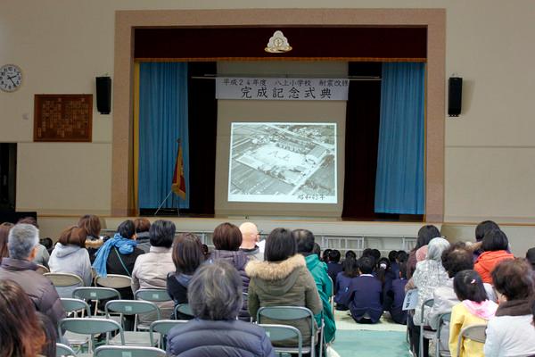 八上小学校の耐震改修完成記念式典にて体育館のステージのスクリーンに八上小学校の白黒写真が映し出されているのを観客や生徒らが見ている写真
