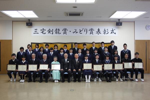 額に入った表彰状を持つ受賞者と、その後ろに立つ学生の集合写真