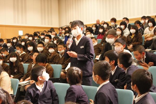マスクを着用した小学生が集まり、1人がマイクを持って挨拶する写真