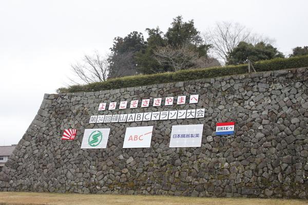 石垣に「ようこそささやまへ」の文字と「第34回篠山ABCマラソン大会」の文字が貼ってある写真