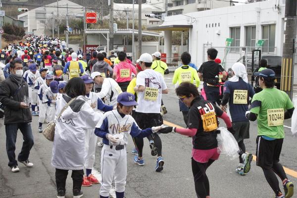 沿道で野球のユニフォームを着た少年たちがマラソンのランナー達にチョコを手渡している写真