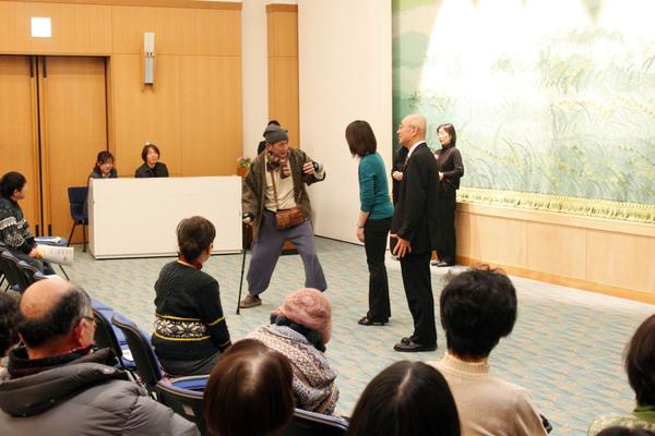 杖を突いて立っている高齢の男性が女性と話しているのを市長が横で見ている様子を観客が見ている写真